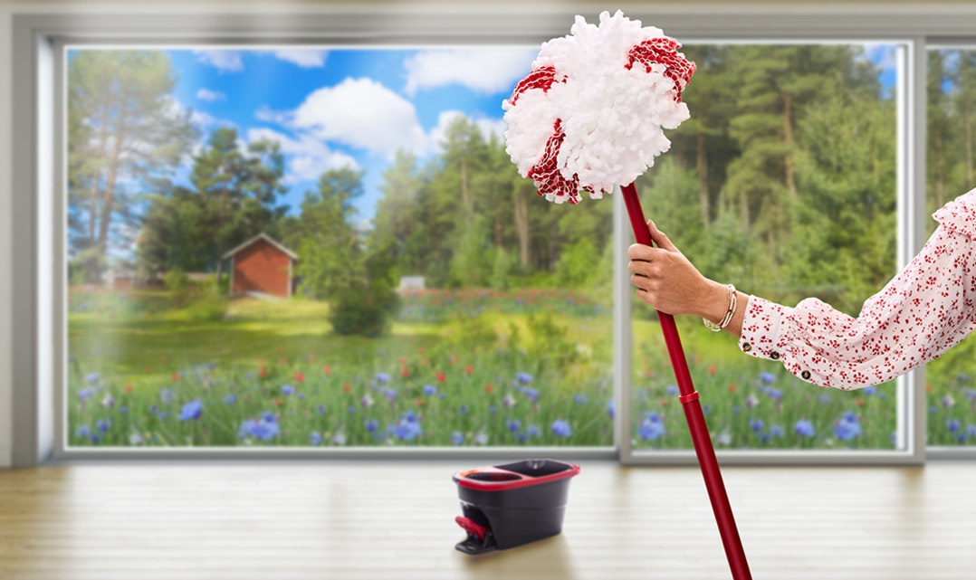 Kesäiseen puseroon pukeutunut käsivarsi pitelee Vileda-moppia kesäniityn näkyessä taustalla.